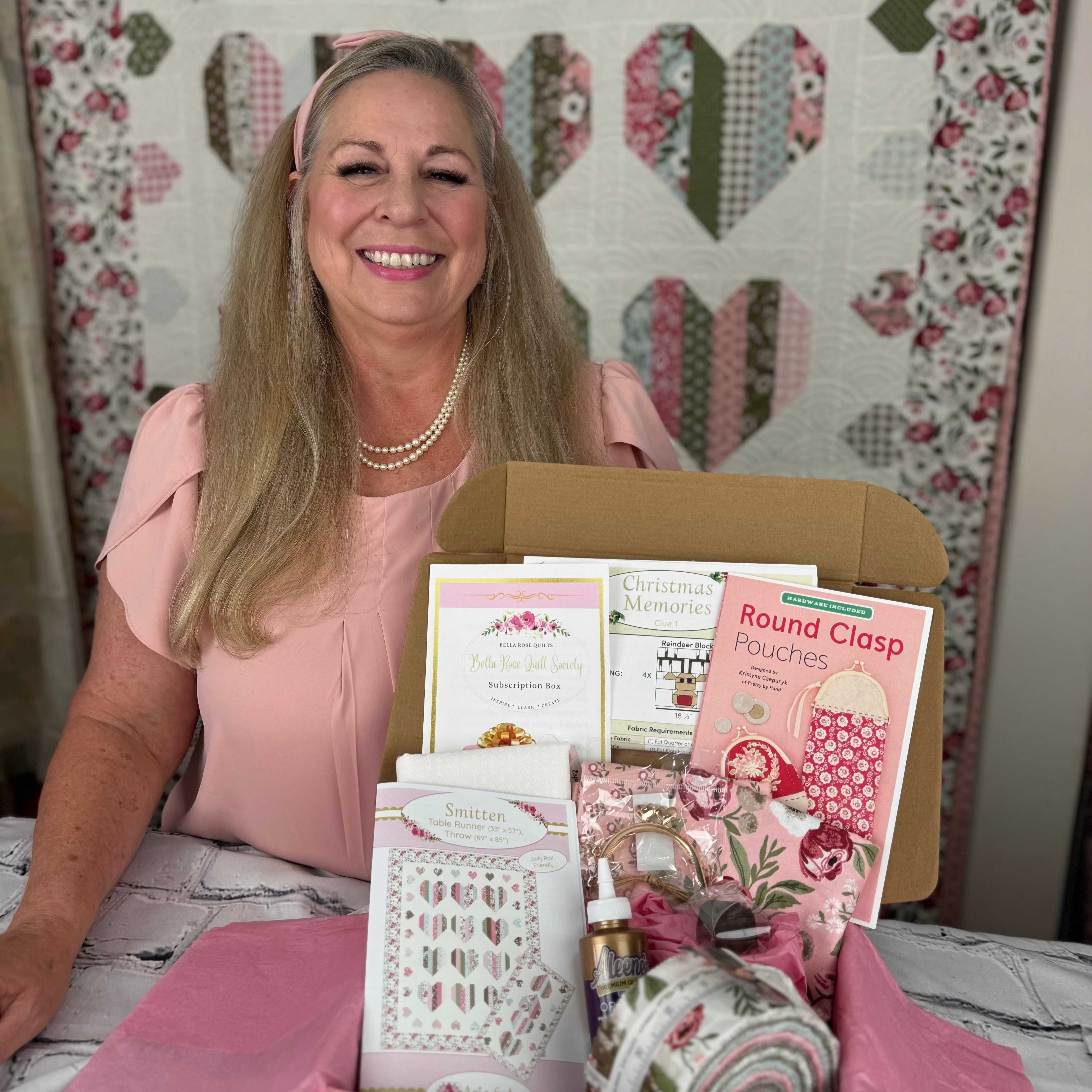 Woman presenting a Bella Rose Quilt Society subscription box with quilting products and patterns on a table in front of a quilt backdrop