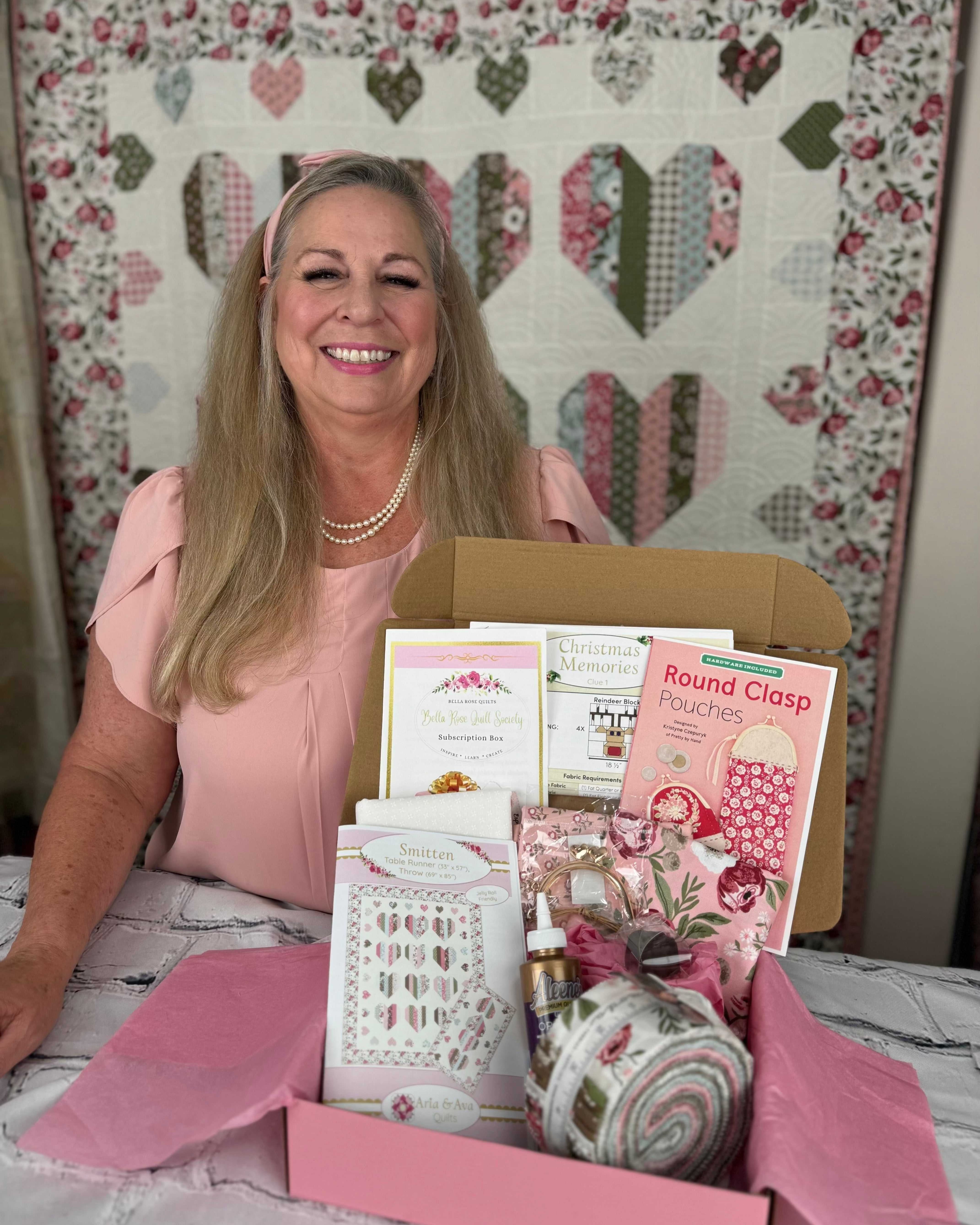 Woman presenting a Bella Rose Quilt Society subscription box with quilting products and patterns on a table in front of a quilt backdrop