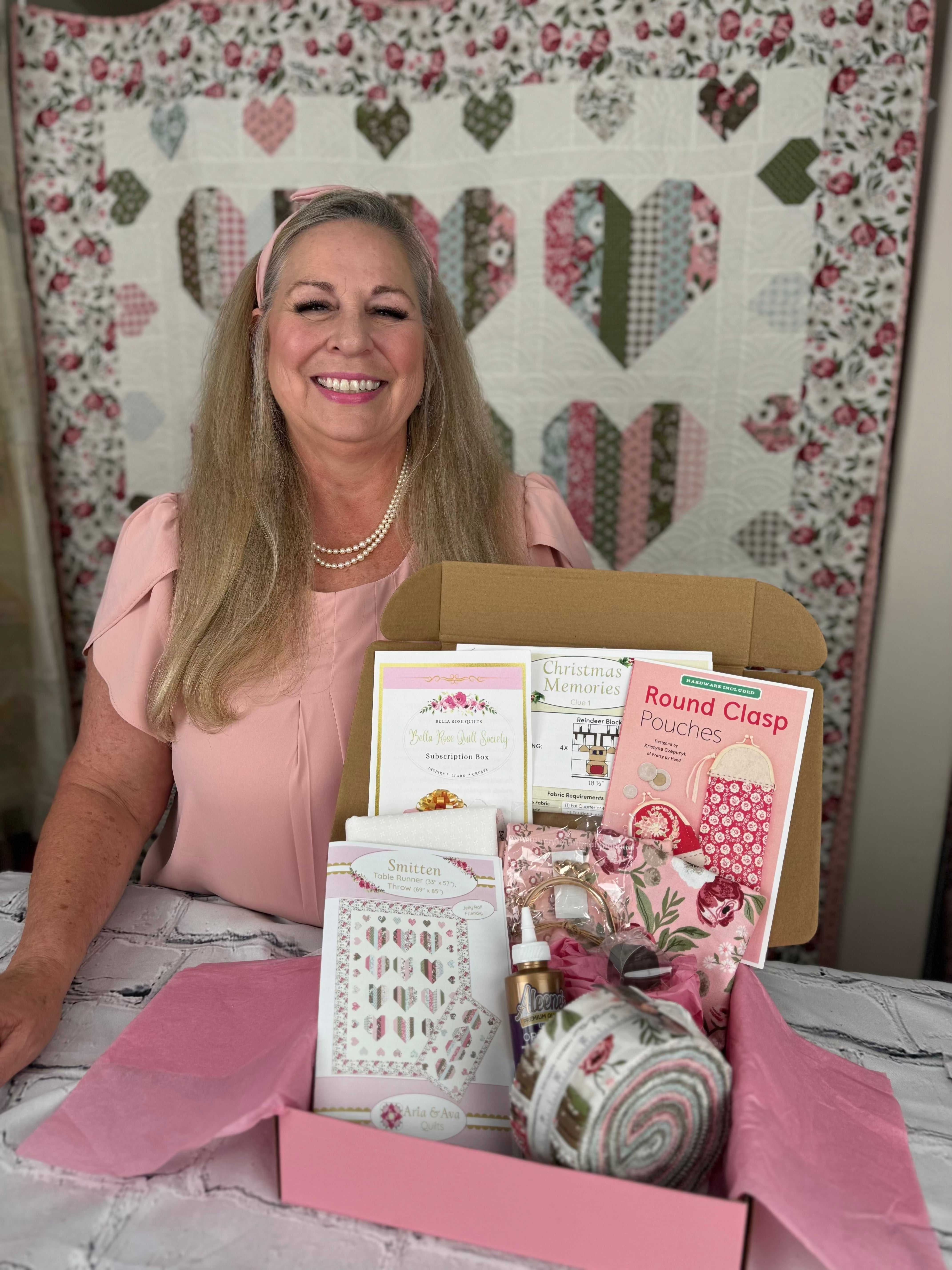 Woman presenting a Bella Rose Quilt Society subscription box with quilting products and patterns on a table in front of a quilt backdrop