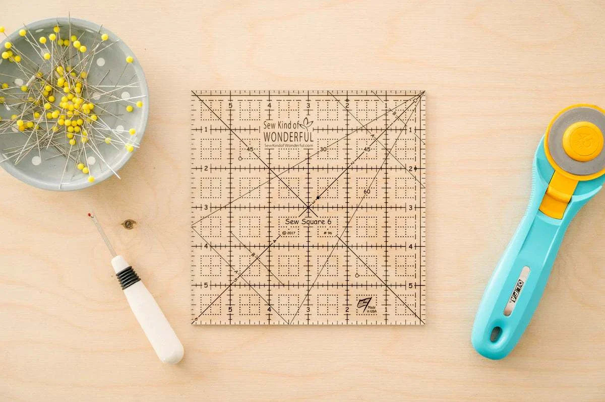 Sew Square 6™ clear acrylic quilting ruler with black grid lines surrounded by sewing tools on wooden surface