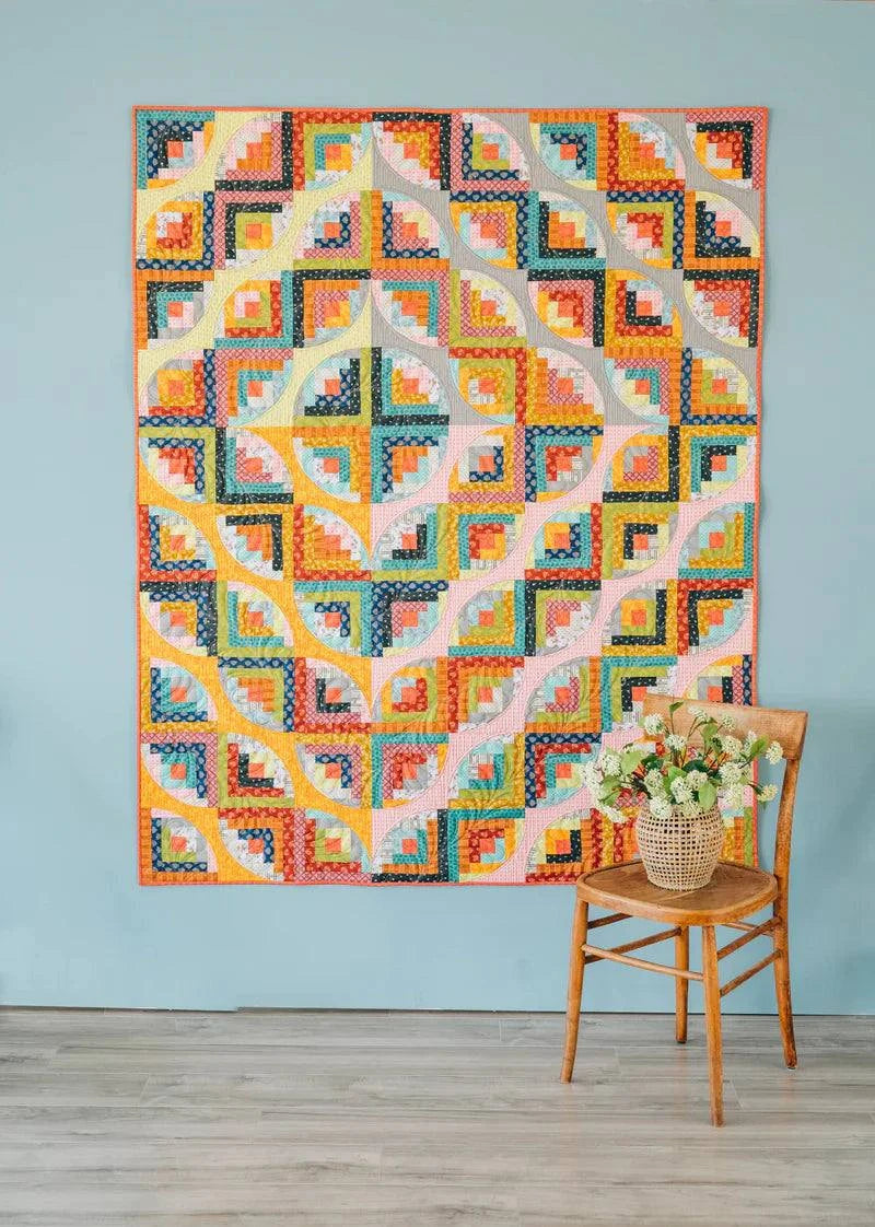 Colorful Curvy Log Cabin quilt with curved patchwork hanging on wall next to wooden chair with flowers