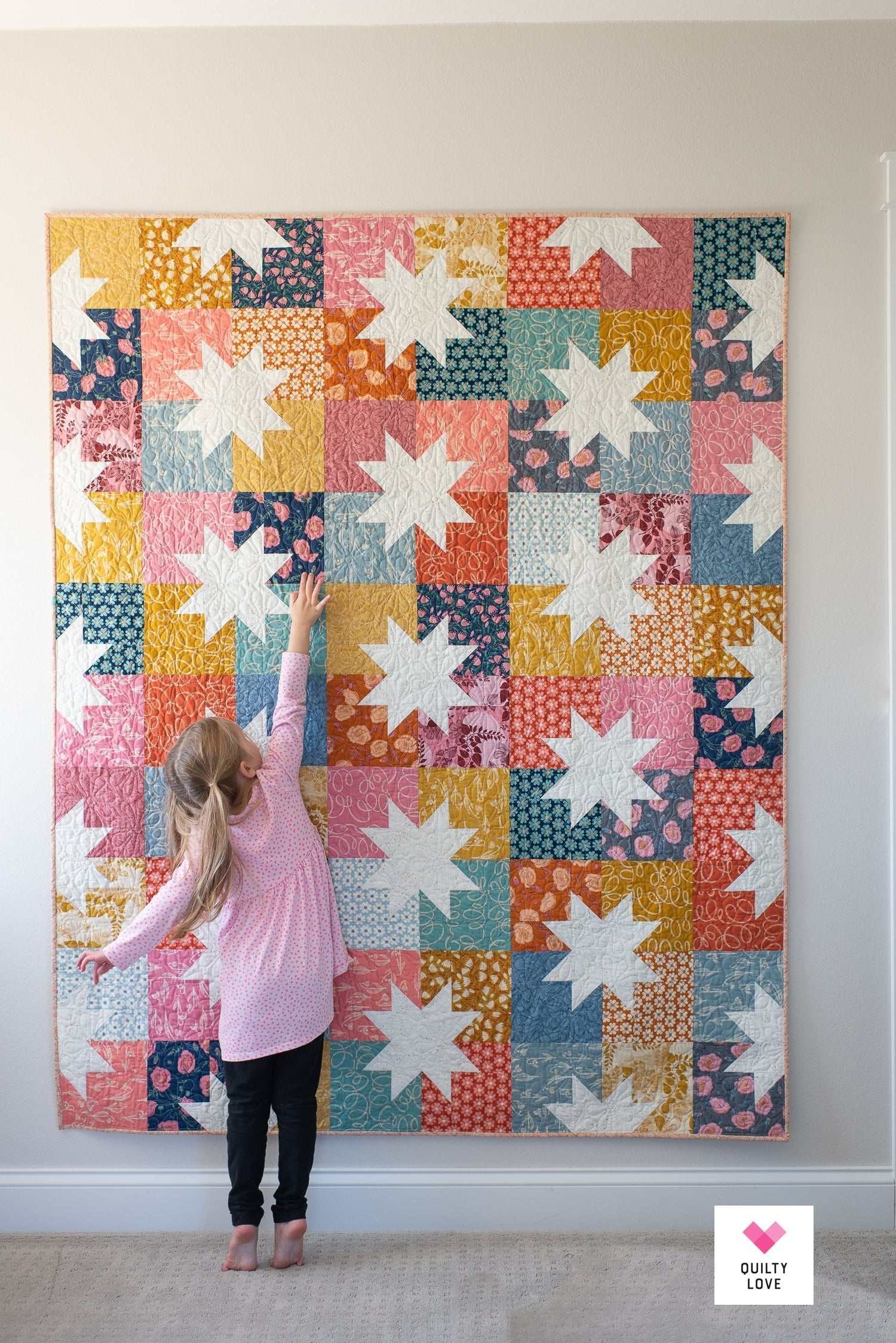 Colorful Star Pop II 2 Quilt Pattern by Quilty Love hanging on wall with child reaching towards it