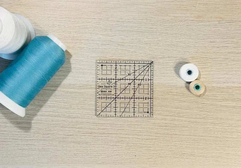 Sew Square 3™ clear 3x3 inch acrylic quilting ruler with black markings and square-up dots next to blue thread spools on wooden surface