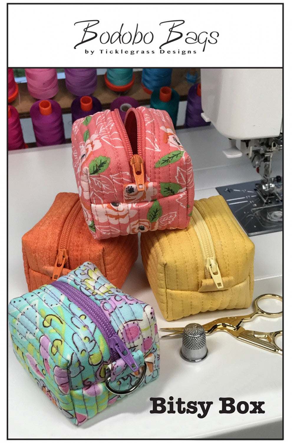 Four colorful small fabric Bitsy Box pouches with zippers by Bodobo Bags next to sewing tools and machine